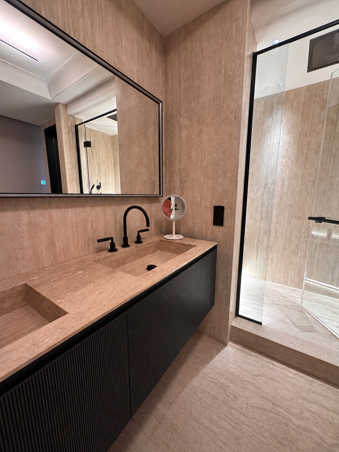 Salle de bains du duplex Maubourg : travertin au sol et aux murs, meuble double vasque noir et douche vitrée